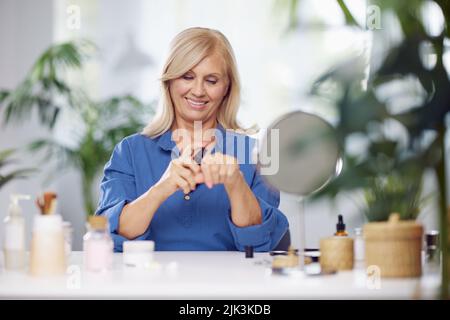 Ritratto di una bella donna anziana che fa il trucco nella camera da letto di fronte allo specchio. Sta mettendo la crema della mano. Foto Stock