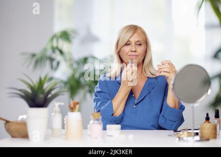 Una bella donna bionda anziana seduta a casa e mettere il rossetto sulle labbra. Foto Stock