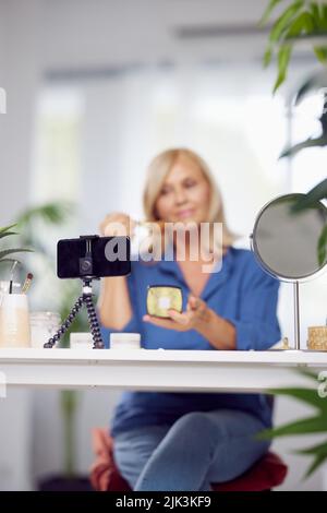 Una bella donna bionda anziana è seduta a casa e fare il suo make-up mattina routine. Donna anziana che mette arrossire il viso Foto Stock