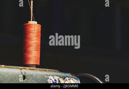 Rocchetto di filo arancione su una vecchia macchina da cucire grigia e sfondo scuro Foto Stock