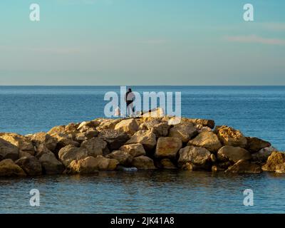 CANNES 02-23-2020 pescatore girato da dietro parla al telefono su un molo di grandi pietre contro il cielo azzurro e il mare al mattino Foto Stock