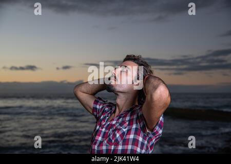 Ragazzo con occhi chiusi e espressione rilassata tocca i capelli mentre si alza in piedi al mare e respira l'aria fresca, concetto di benessere. Foto Stock