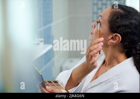 La giovane donna stupefacente tiene una tavolozza colorata dell'ombretto e usando un pennello per il make-up, applica l'ombra dell'occhio alla palpebra superiore. Foto Stock