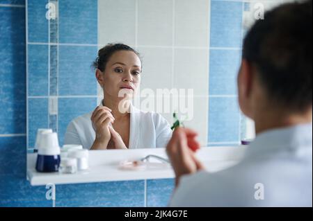 Riflesso nello specchio del bagno di una donna serena massaggiante le mani imbevute di idratante, curando la pelle delle mani Foto Stock