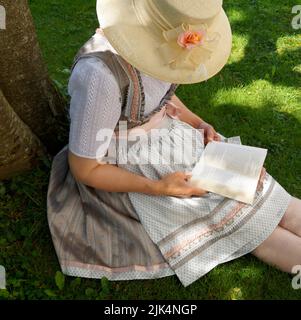 Una donna in un bel vestito tradizionale bavarese dirndl (o Tracht) leggendo un libro sul prato verde in una giornata estiva soleggiata (Monaco, Baviera, tedesco Foto Stock