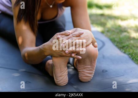 Primo piano della donna seduta in una posa di yoga curva in avanti. Una signora allunga i piedi e i tendini con le mani nella posa del paschimottanasana yoga. Foto Stock