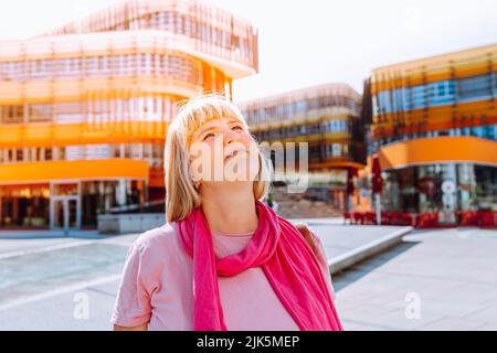 donna bionda di mezza età 40s con capelli lunghi, in sciarpa rosa e abiti estivi, guarda all'architettura moderna con speranza e sognamento. concetto di husi Foto Stock