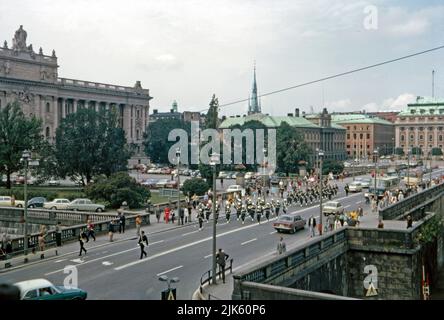 Stoccolma nel 1970 – una vista dall'angolo tra Norrström e Slottskajan del Parlamento (Riksdagshuset) sulla sinistra. Una banda militare sta marciando verso Norrström. L'edificio in stile neoclassico, progettato da Aron Johansson, si trova sul sito dell'isola di Helgeandsholmen. Le auto parcheggiate di fronte all'edificio sono un ricordo del passato dato che l'area è ora una piazza ben curata e l'area parcheggio erbosa. L'edificio dietro (centro a destra) è il Dipartimento degli affari Esteri (Utrikesdepartementet Sverige), una fotografia d'epoca degli anni '1970. Foto Stock