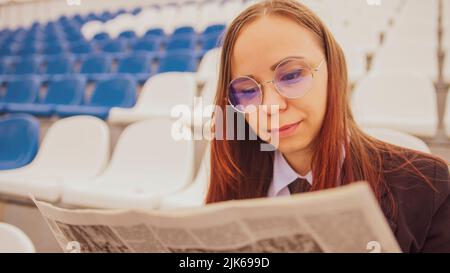 Una donna in occhiali e un abito da lavoro legge un giornale mentre si siede allo stadio. Foto Stock