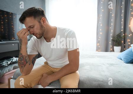 L'uomo giovane e stanco siede in una camera su un letto confortevole Foto Stock