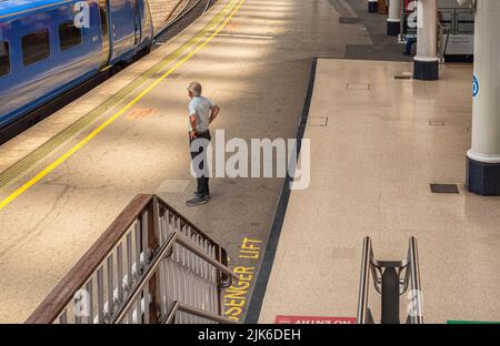 Un uomo è in piedi su una piattaforma della stazione ferroviaria guardando una velocità del treno vicino. I corrimano sono in primo piano. Foto Stock