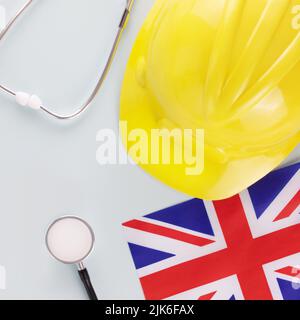 Assistenza medica e assicurazione dei lavoratori in Gran Bretagna. Bandiera della Gran Bretagna, stetoscopio e hardhat giallo su sfondo blu. Sicurezza del lavoro e r Foto Stock