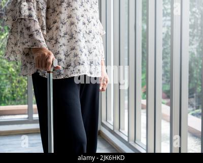 Primo piano la mano della donna anziana che tiene una canna da passeggio in piedi da sola vicino alla finestra di vetro nella stanza con spazio copia. Donna anziana solitaria, il grandmo Foto Stock