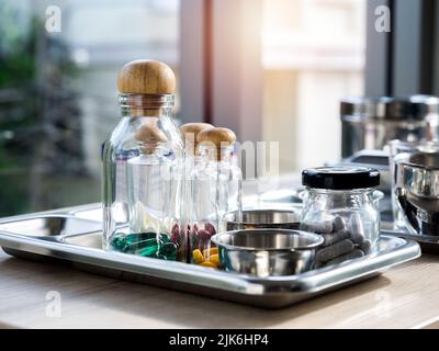 Primo piano set di vitamine e integratori nelle piccole bottiglie di vetro trasparente nel vassoio medico in acciaio su tavola di legno. Vitamina verde, gialla, rossa, bianca Foto Stock