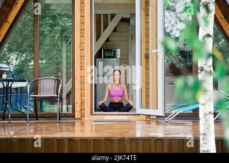 La donna si siede in posizione di yoga lotus in una casa forestale di fronte alla porta aperta, godendo di aria fresca dopo la pioggia Foto Stock