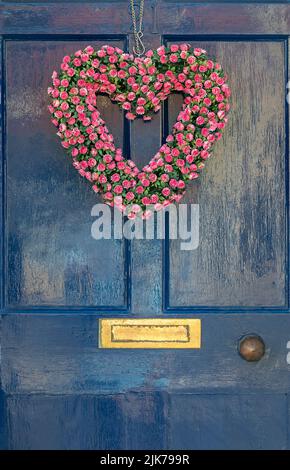 porta frontale in legno dipinto di blu con una corona fatta di fiori rosa a forma di cuore e mobili in ottone, porta sospesa a forma di cuore Foto Stock