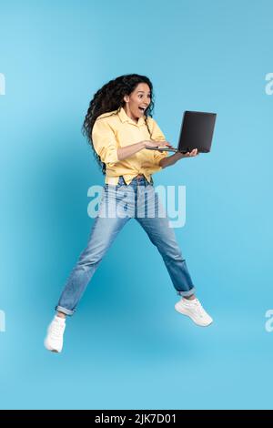 Eccitati laptop Lady Holding mediorientali che saltano su sfondo blu Foto Stock