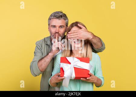 L'uomo maturo ha ricevuto sorpresa per la sua moglie amorevole, donna che tiene la scatola del regalo, l'uomo che copre gli occhi e che mostra il gesto di silenzio Foto Stock