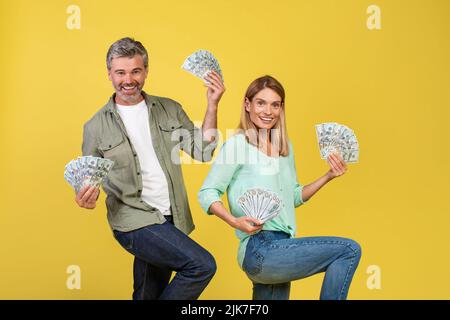 Coppia matura eccitata ballando con un mazzo di banconote in denaro e celebrando il successo su sfondo giallo studio Foto Stock
