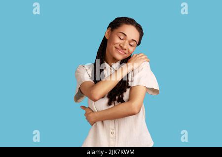 Giovane donna allegra con cialde nere sorride teneramente, tocca le spalle, si abbraccia, tiene gli occhi chiusi, indossa una camicia bianca. Studio interno girato isolato su sfondo blu. Foto Stock