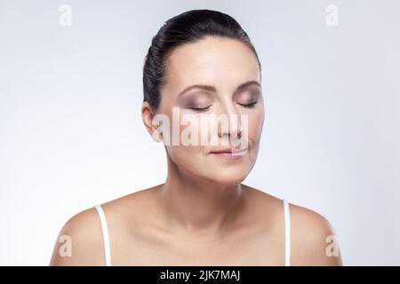 Primo piano ritratto di bella donna di mezza età con pelle perfetta sorridente dopo crema, balsamo, maschera, facendo ringiovanimento procedure, mantiene gli occhi chiusi. Studio interno girato isolato su sfondo bianco. Foto Stock