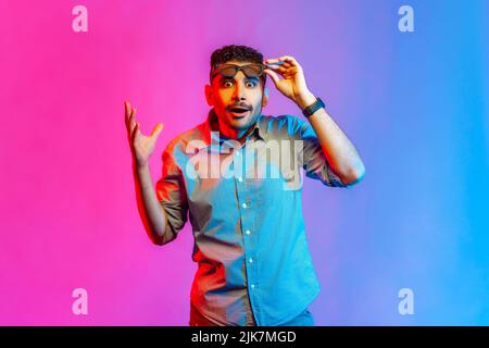 Ritratto di uomo sorpreso in camicia in piedi occhiali e mano rialzati, guardando la macchina fotografica con occhi grandi ed espressione scioccata. Studio interno girato isolato su colorato sfondo di luce al neon. Foto Stock