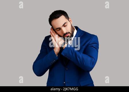 Ritratto di uomo stanco che dorme sulle palme, tenendo gli occhi chiusi, indossando un maglione bianco, indossando una tuta ufficiale. Studio interno girato isolato su sfondo grigio. Foto Stock