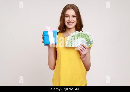 Shopping di regali. Ragazza adolescente felice in T-shirt gialla con cassa attuale e banconote in euro, soddisfatto con acquisto, rimborso e prestito bancario. Studio interno girato isolato su sfondo grigio. Foto Stock