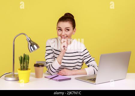 Donna felice dipendente sul posto di lavoro che si arrossiva con un gesto di silenzio, tenendo il dito sulle labbra e sorridendo alla fotocamera, chiedendo di tenere segreto. Studio interno girato isolato su sfondo giallo. Foto Stock