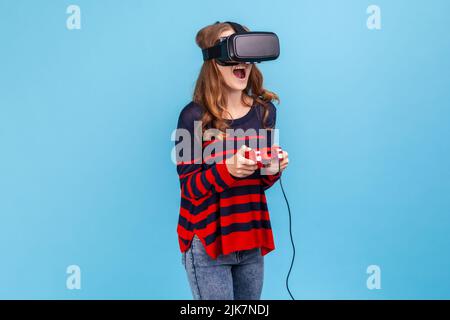 Donna stupita che indossa un pullover casual a righe e un visore vr, giocando a videogame per realtà virtuale con joypad, ha emozionato il sorriso sul suo volto. Studio interno girato isolato su sfondo blu. Foto Stock