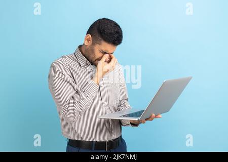 Ritratto di giovane uomo d'affari adulto in piedi con il laptop in mani, strofinando gli occhi, sembra stanco e esausto, indossando una camicia a righe. Studio interno girato isolato su sfondo blu. Foto Stock