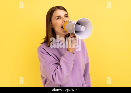 Ritratto di bella donna urlando forte in megafono, annunciando importante pubblicità, protestando, indossando felpa con cappuccio viola. Studio interno girato isolato su sfondo giallo. Foto Stock