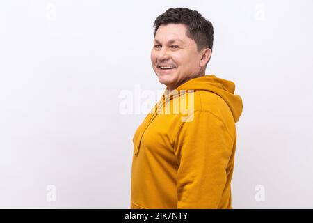 Vista laterale di un bell'uomo di mezza età con capelli scuri in piedi guardando la fotocamera con il volto soddisfatto e sorridente, indossando una felpa con cappuccio urban style. Studio interno girato isolato su sfondo bianco. Foto Stock