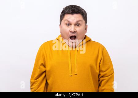 Ritratto di uomo eccitato con bocca aperta e grandi occhi stupiti, guardando sorpreso e sciocco alla macchina fotografica, meravigliato espressione, indossando una felpa con cappuccio urban style. Studio interno girato isolato su sfondo bianco. Foto Stock