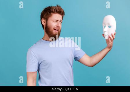 Ritratto di confuso puzzled bel giovane adulto ported uomo che tiene la maschera bianca nelle mani e guardando con la bocca aperta, disprezza la personalità nascosta. Studio interno girato isolato su sfondo blu. Foto Stock