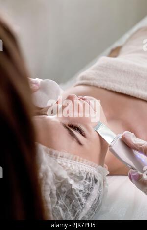 Cosmetologist, estetician che fa il trattamento facciale con la spatola ultrasonica alla donna giovane, trattamento di scrubber della pelle del viso con la spatola ultrasonica, procedura di pulizia facciale nel salone di bellezza. Foto Stock