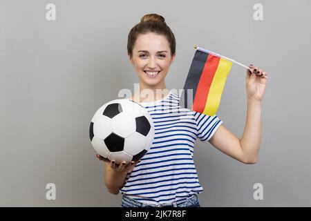 Ritratto di gioiosa donna allegra indossando T-shirt a righe con palla di calcio e bandiera tedesca, guardando sorridendo la macchina fotografica, allegro. Studio interno girato isolato su sfondo grigio. Foto Stock