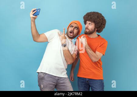 Ritratto di divertente cool due giovani adulti hipster uomini blogger che trasmettono in live streaming o prendere selfie, mostrando rock and roll e shaka gesti. Studio interno girato isolato su sfondo blu. Foto Stock