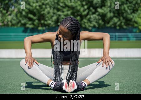 La donna afroamericana si siede sulla pista verde a terra di sport che allunga le gambe nella giornata estiva di sole Foto Stock