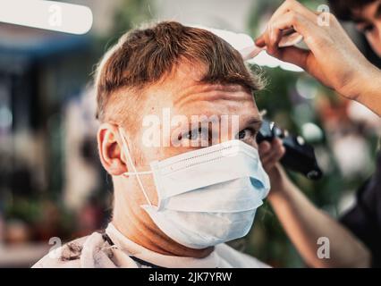Salone fotografico Lifestyle. Il cliente giovane indossa la maschera facciale, lo sguardo sorpreso insoddisfatto, la fronte rugosa. Pettine da lavoro a mano maschio per parrucchieri Foto Stock