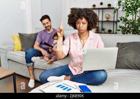 Madre di famiglia interracial che lavora usando computer portatile a casa puntando verso l'alto guardando triste e sconvolto, indicando direzione con le dita, infelice e d Foto Stock