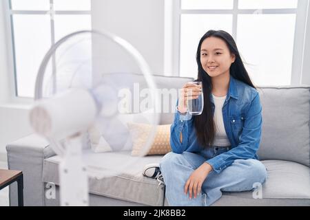 Giovane donna asiatica che beve un bicchiere d'acqua godendo l'aria dal ventilatore che osserva positivo e felice in piedi e sorridendo con un sorriso sicuro che mostra i denti Foto Stock