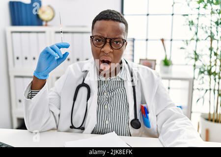 Giovane medico africano che tiene la siringa all'ospedale in faccia shock, guardando scettico e sarcastico, sorpreso con bocca aperta Foto Stock