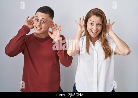 Madre e figlio si alzano insieme su un fondo isolato cercando di sentire entrambe le mani sull'orecchio gesto, curioso per pettegolezzi. Problema uditivo, sordi Foto Stock