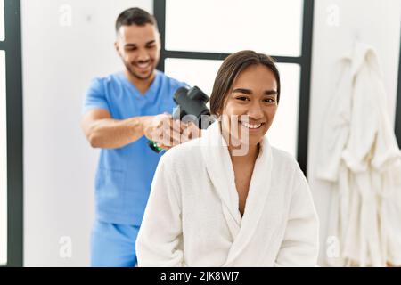 Latino uomo e donna che indossano la fisioterapia uniforme massaggiante collo con pistola a percussione al centro di bellezza Foto Stock