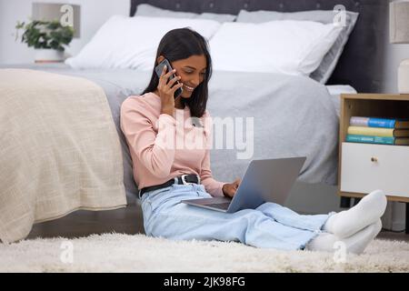 Grazie per avermi chiamato. Scatto a tutta lunghezza di una giovane donna attraente che fa una telefonata mentre usando il suo laptop nella camera da letto a casa. Foto Stock