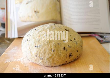 una palla di pasta artigianale per pane su un pannello di legno con un libro di ricette sfocato sullo sfondo Foto Stock