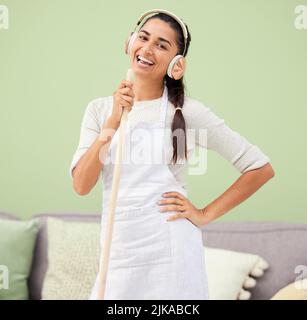Ive ha acceso la mia musica. È il momento di pulire. Una giovane donna attraente che ascolta musica e canta mentre usa una scopa per spazzare la sua casa. Foto Stock