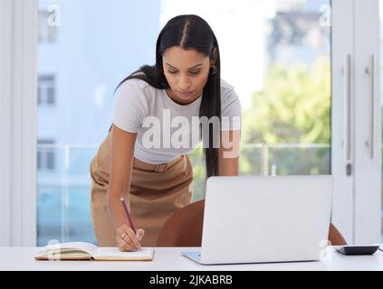 Il mio programma è pieno di riunioni. Una giovane donna d'affari attraente in piedi da sola sulla sua scrivania in ufficio e scrivere nel suo notebook. Foto Stock