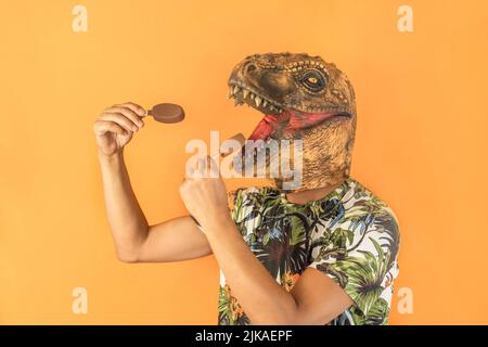 Uomo in dinosauro animale testa maschera mangiare due gelato al cioccolato isolato su sfondo giallo con spazio copia Foto Stock